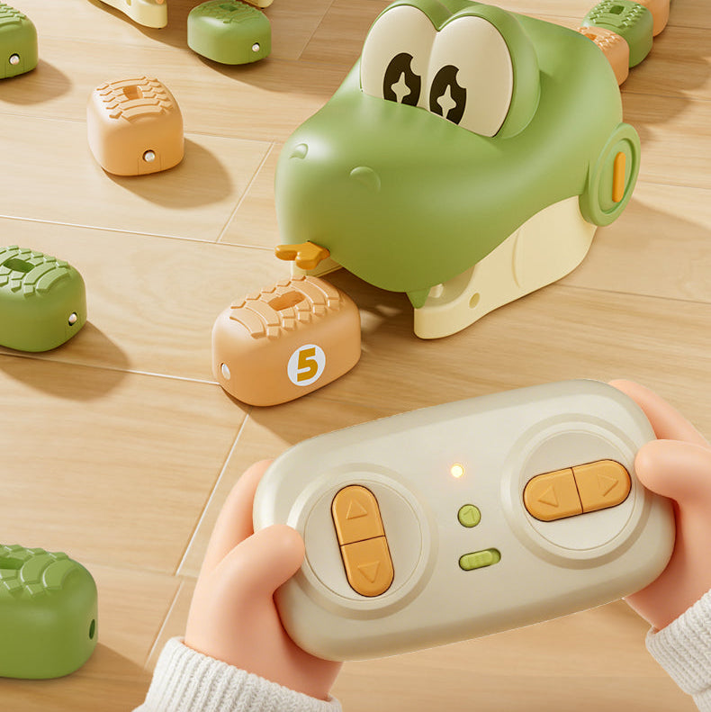 A child's hands hold a light gray remote control with orange buttons, pointing it at a green crocodile-shaped toy that is picking up a tan numbered block on a light wood floor. More toy blocks are scattered around.