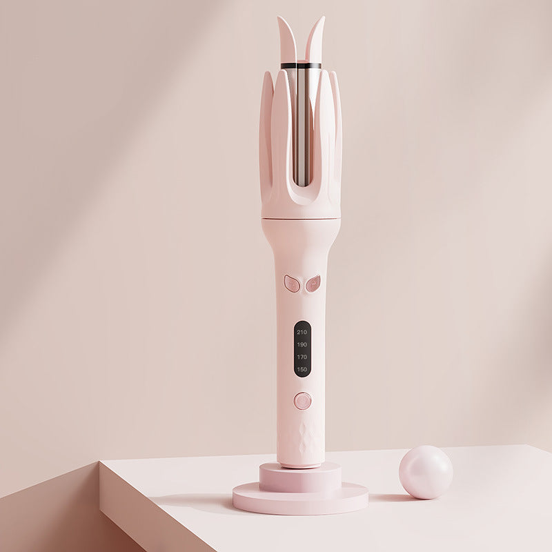 A pink hair styling tool with a curved handle, dual-pronged barrel, and small digital display, shown standing on a matching pink stand. The background is a soft beige studio setting with a small white ball prop.