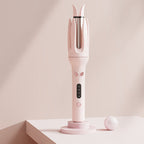 A pink hair styling tool with a curved handle, dual-pronged barrel, and small digital display, shown standing on a matching pink stand. The background is a soft beige studio setting with a small white ball prop.
